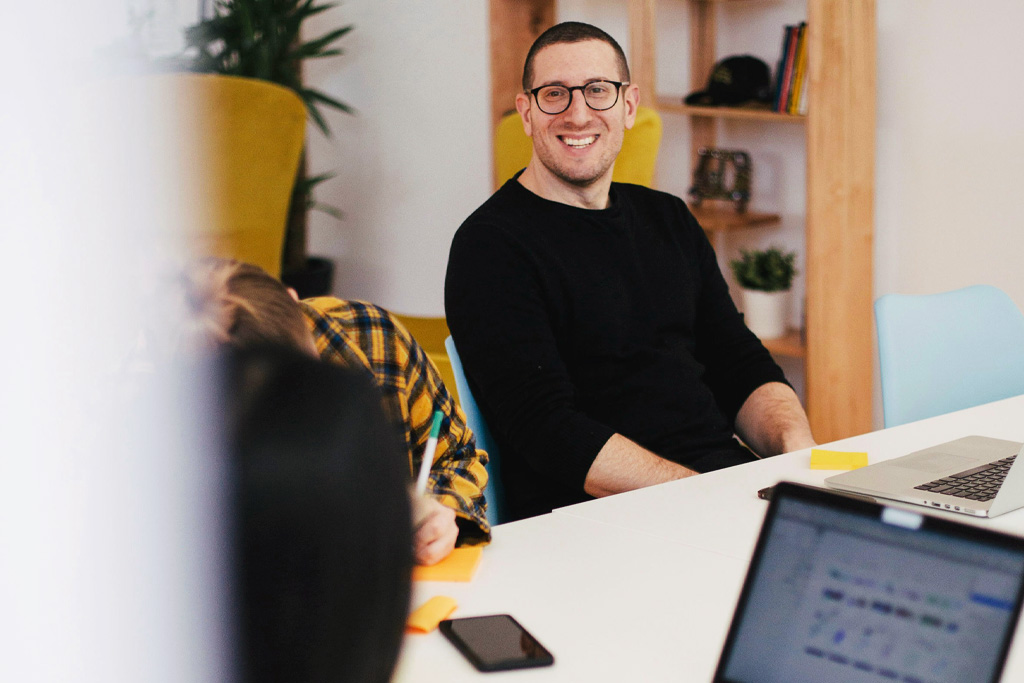 Studenter sitter och pratar runt ett bord. Det är något slags möte med laptops, anteckningsblock och penna. Ser glada ut.