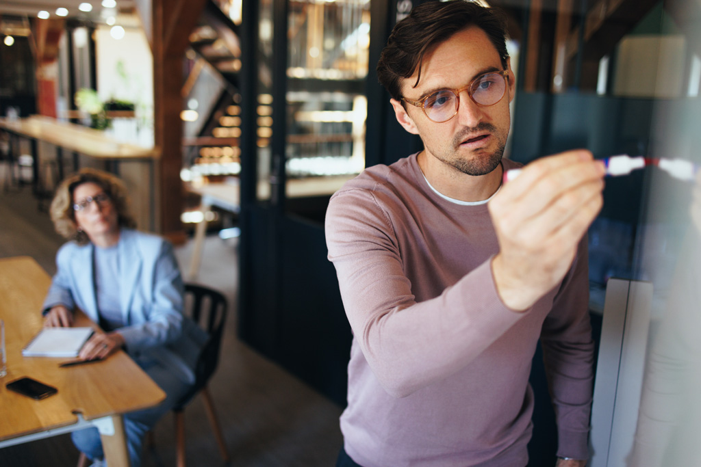En revisor skriver på en whiteboardtavla. Informerar en åhörare om krav och regler för certifiering.