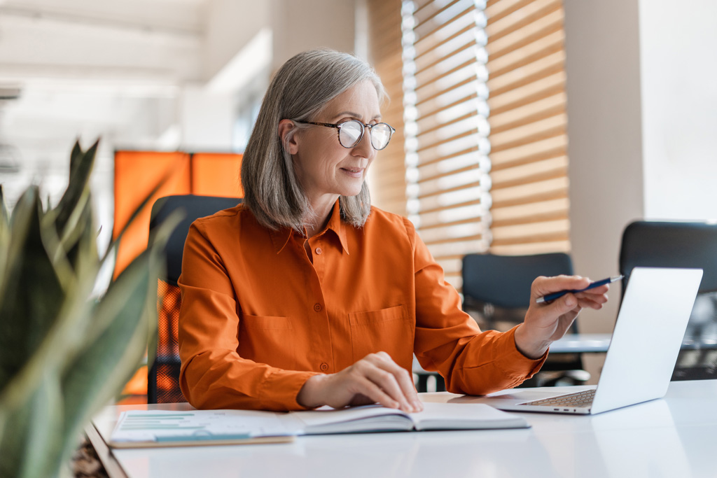 En senior revisor, med färgglad orange blus, arbetar med sin dator.