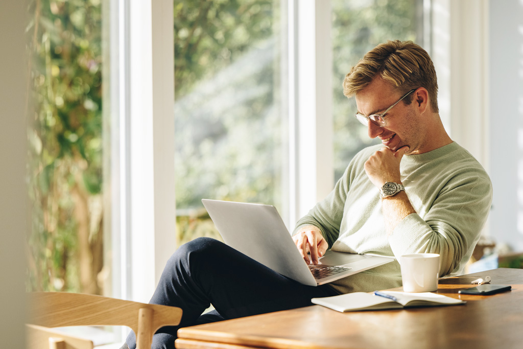 Person sitter vid ett lunchbord på jobbet. Stora fönster i bakgrunden släpper in ett varmt ljus. Mannen går en online-kurs i kommunal räkenskapsrevision.